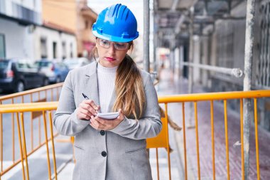 Young beautiful hispanic woman architect writing on notebook at street