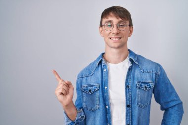Caucasian blond man standing wearing glasses with a big smile on face, pointing with hand finger to the side looking at the camera. 