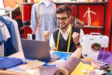 Dövmeli İspanyol genç adam terzi, terzi odasında dizüstü bilgisayar kullanıyor. Gururla bağırıyor, zaferi kutluyor ve başarıyı kollarını kaldırarak kutluyor. 