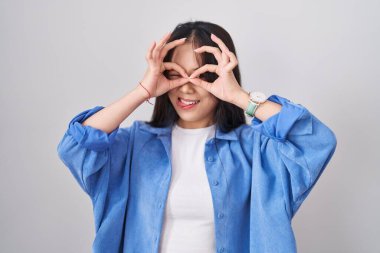 Young chinese woman standing over white background doing ok gesture like binoculars sticking tongue out, eyes looking through fingers. crazy expression. 