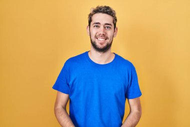 Hispanic man with beard standing over yellow background with hands together and crossed fingers smiling relaxed and cheerful. success and optimistic 