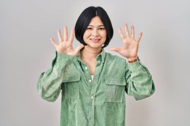 Young asian woman standing over white background showing and pointing up with fingers number ten while smiling confident and happy. 