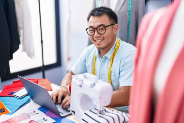 Young chinese man tailor smiling confident using laptop at tailor shop