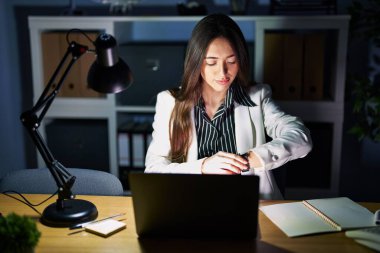 Genç esmer kadın geceleri ofiste dizüstü bilgisayarla çalışıyor. Saati kontrol ediyor, rahat ve kendinden emin. 