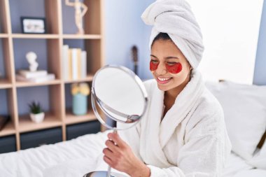 Yatak odasındaki aynaya bakan genç Latin kadın dolgun göz tedavisi görüyor.