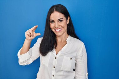 Mavi arka planda duran, gülümseyen, kendine güvenen, el hareketi yapan, parmakları küçük boy işareti yapan ve kameraya bakan genç İspanyol kadın. ölçüm kavramı. 
