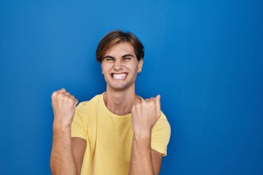 Young man standing over blue background very happy and excited doing winner gesture with arms raised, smiling and screaming for success. celebration concept. 