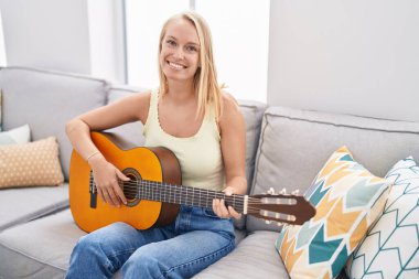 Genç sarışın kadın evdeki kanepede oturmuş klasik gitar çalıyor.