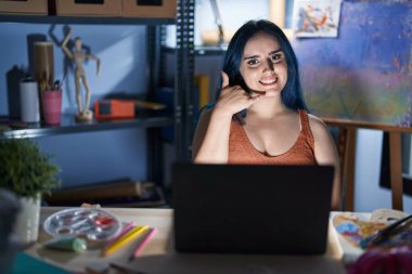 Young modern girl with blue hair sitting at art studio with laptop at night smiling doing phone gesture with hand and fingers like talking on the telephone. communicating concepts. 