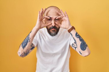 Sarı arka planda duran İspanyol dövmeli genç adam parmaklarıyla gözlerini açmaya çalışıyor, uykulu ve yorgun bir şekilde sabah yorgunluğu çekiyor. 