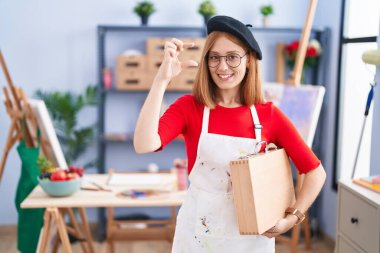 Young redhead woman at art studio holding art case smiling and confident gesturing with hand doing small size sign with fingers looking and the camera. measure concept. 