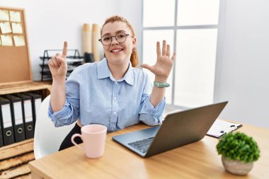 Ofiste çalışan genç kızıl saçlı kadın bilgisayarını kullanarak altı numaralı parmaklarını gösterip gülümseyerek mutlu ve kendinden emin bir şekilde gülümsüyor.. 