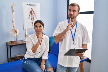Young hispanic woman at physiotherapist appointment thinking concentrated about doubt with finger on chin and looking up wondering 