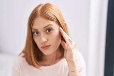 Young redhead woman touching face sitting on bed at bedroom