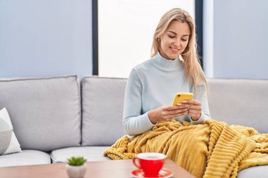 Evdeki kanepede oturan akıllı telefon kullanan genç sarışın kadın.