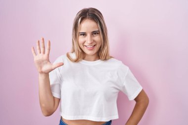 Pembe arka planda duran genç sarışın kadın beş numarayı gösterip işaret ederken kendinden emin ve mutlu bir şekilde gülümsüyor.. 