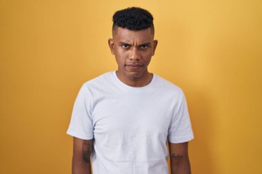 Sarı arka planda duran Latin asıllı genç adam şüpheci ve gergin, sorun yüzünden surat asıyor. negatif kişi. 