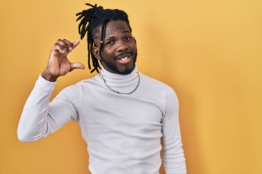 Sarı arka planda balıkçı yaka kazak giyen Afrikalı bir adam gülümsüyor ve el hareketleriyle küçük boy işareti yapıyor, parmaklarıyla bakıyor ve kameraya bakıyor. ölçüm kavramı. 