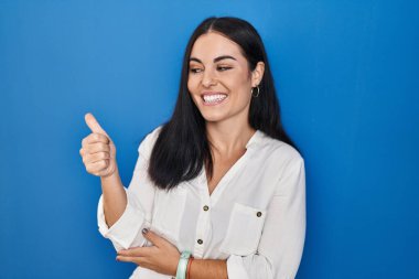 Young hispanic woman standing over blue background looking proud, smiling doing thumbs up gesture to the side 