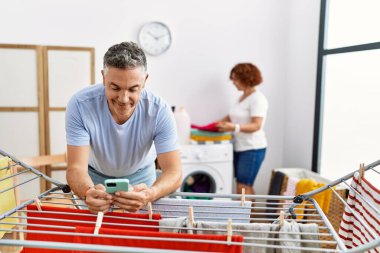Orta yaşlı erkek ve kadın çamaşırhanede çamaşır asan akıllı telefon kullanan bir çift.
