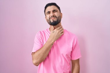 İspanyol genç adam pembe arka planda duruyor. Ağrılı boyuna dokunuyor. Grip, mantar ve enfeksiyon yüzünden boğazı ağrıyor. 