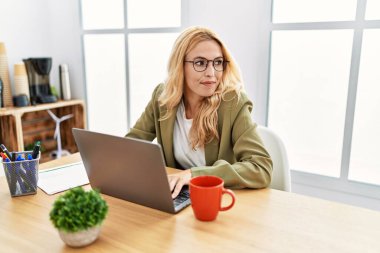 Beautiful blonde woman working at the office with laptop looking to side, relax profile pose with natural face with confident smile. 