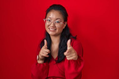 Asian young woman standing over red background pointing fingers to camera with happy and funny face. good energy and vibes. 