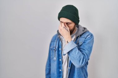 Dövmeleri olan, yün şapkalı, burnu ve gözleri ovuşturmaktan yorgun, yorgun ve başı ağrıyan genç İspanyol bir adam. Stres ve hüsran kavramı. 