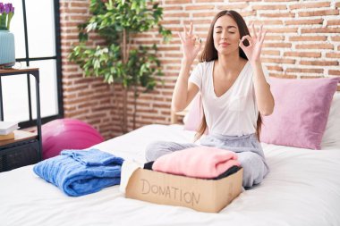 Genç İspanyol kadın, kullanılmış giysileri bağış kutusuna koyuyor yatağa oturuyor, rahatlıyor ve gözleri kapalı gülümsüyor parmaklarıyla meditasyon hareketi yapıyor. yoga konsepti. 