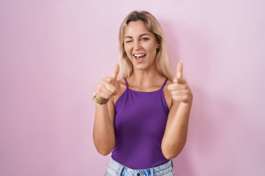 Young blonde woman standing over pink background pointing fingers to camera with happy and funny face. good energy and vibes. 