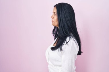 Olgun İspanyol kadın pembe arka planda duruyor. Yan tarafa bakıyor, rahat poz veriyor ve kendine güvenen bir gülümseme sergiliyor.. 