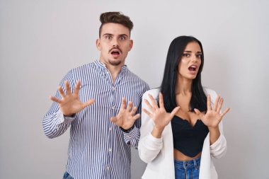 Young hispanic couple standing over white background afraid and terrified with fear expression stop gesture with hands, shouting in shock. panic concept. 