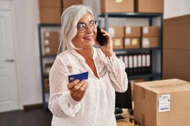 Orta yaşlı kadın ecommerce iş işçisi. Ofiste kredi kartıyla akıllı telefondan konuşuyor.