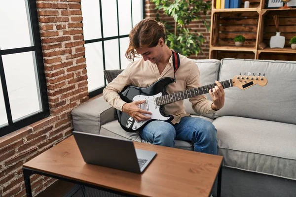 Young caucasian man having online electrical guitar class sitting on sofa at home