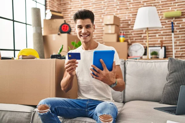 Young hispanic man using touchpad and credit card sitting on sofa at new home