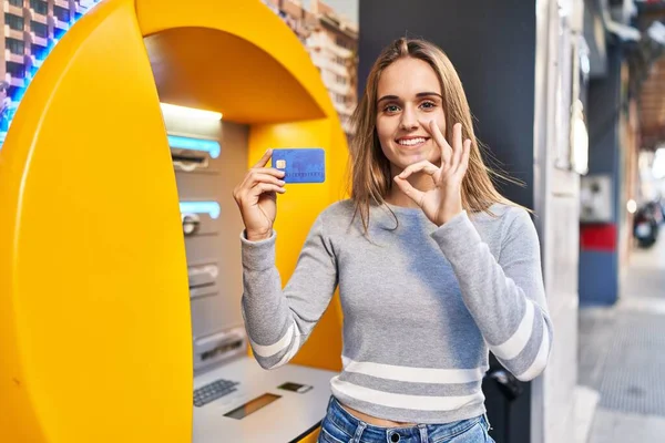 Young doctor woman holding credit card at cash point doing ok sign with fingers, smiling friendly gesturing excellent symbol