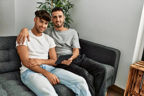 Two hispanic men couple using touchpad hugging each other at home