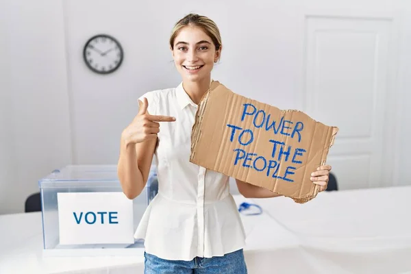 Young blonde woman at political election holding power to the people banner smiling happy pointing with hand and finger