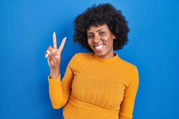 Black woman with curly hair standing over blue background smiling looking to the camera showing fingers doing victory sign. number two.