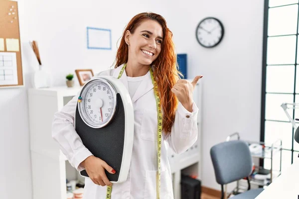 Young Redhead Woman Nutritionist Doctor Holding Weighing Machine Pointing Thumb — Stock Photo, Image