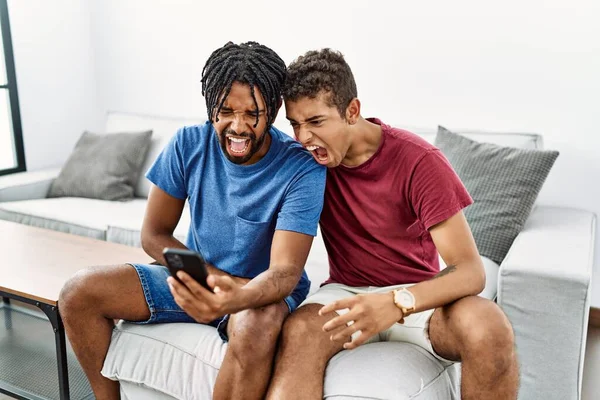Young hispanic men using smartphone sitting on the sofa at home angry and mad screaming frustrated and furious, shouting with anger. rage and aggressive concept.