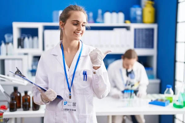 Blonde woman working at scientist laboratory pointing thumb up to the side smiling happy with open mouth