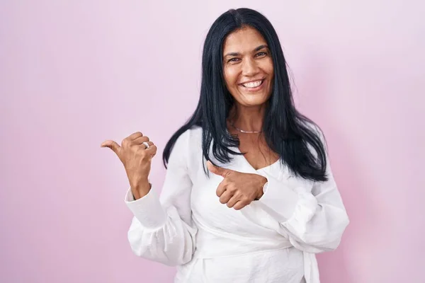 Mature hispanic woman standing over pink background pointing to the back behind with hand and thumbs up, smiling confident