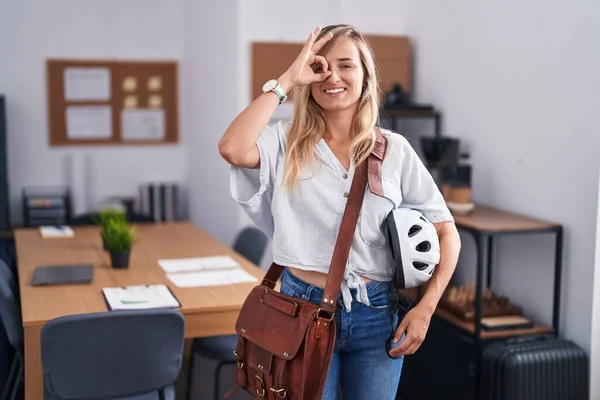 Young blonde woman working at the office holding bike helmet smiling happy doing ok sign with hand on eye looking through fingers