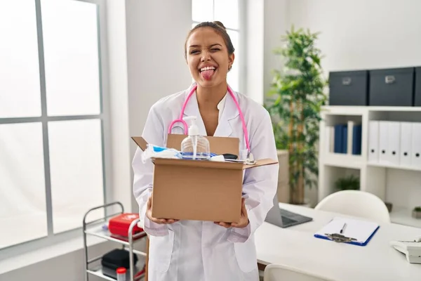 Young Hispanic Doctor Holding Box Medical Items Sticking Tongue Out — Stock Photo, Image