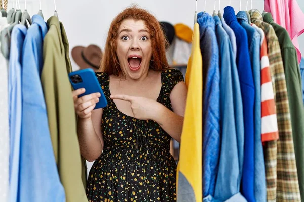 Genç kızıl saçlı kadın elbise rafında kıyafet arıyor. Akıllı telefon kullanıyor. Gülümsüyor. 