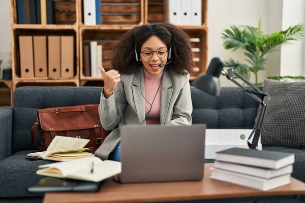 Young african american woman doing online session at consultation office pointing thumb up to the side smiling happy with open mouth 