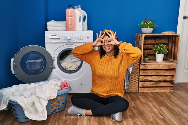 Young hispanic woman doing laundry doing ok gesture like binoculars sticking tongue out, eyes looking through fingers. crazy expression.