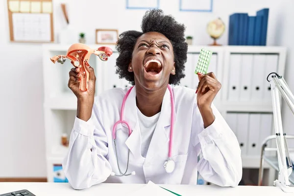 African doctor woman holding anatomical female genital organ and birth control pills angry and mad screaming frustrated and furious, shouting with anger looking up.