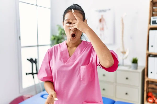 Young hispanic woman working at pain recovery clinic peeking in shock covering face and eyes with hand, looking through fingers with embarrassed expression.